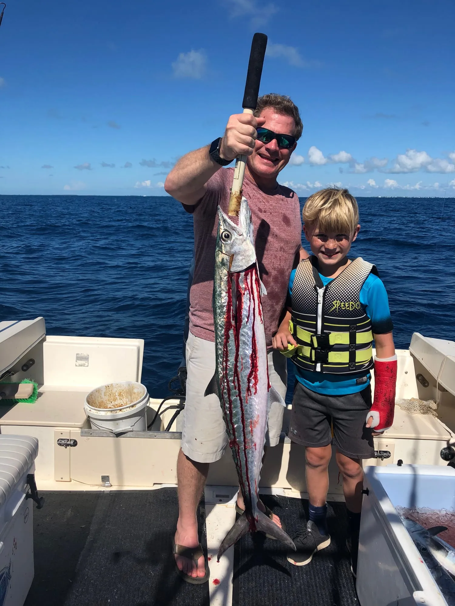 Young boy in a life jacket next to a big catch with his dad